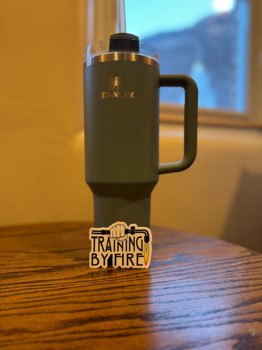 A sticker with a jeweler’s torch design and the phrase 'Training By Fire' displayed on a wooden surface.