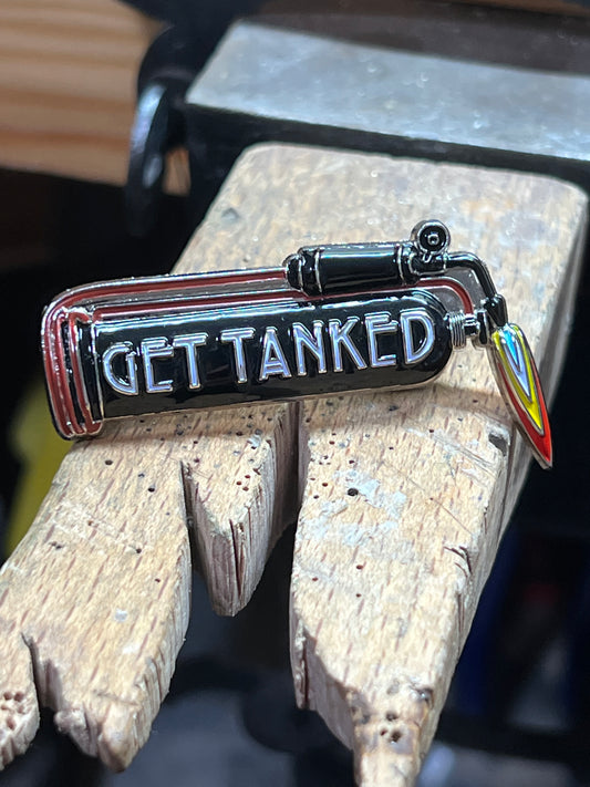 "Get Tanked" enamel pin – front view of hard enamel black and silver pickle tank pin with flame charm on wooden surface