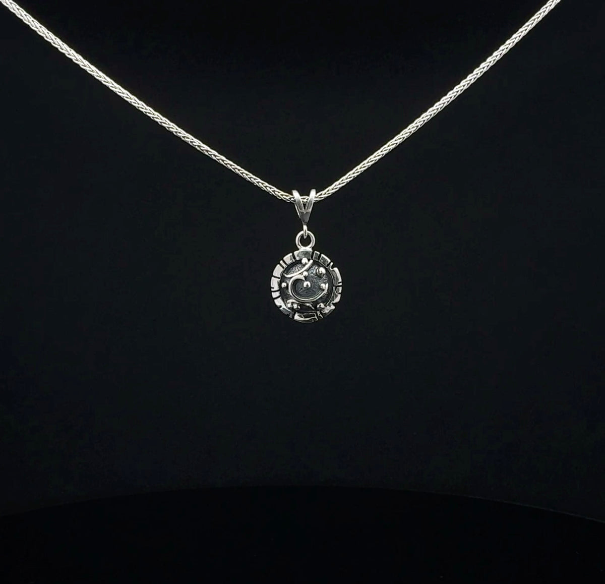A silver necklace with a small circular pendant featuring a stylized face, displayed against a solid black background.