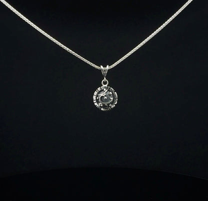 A silver necklace with a small circular pendant featuring a stylized face, displayed against a solid black background.