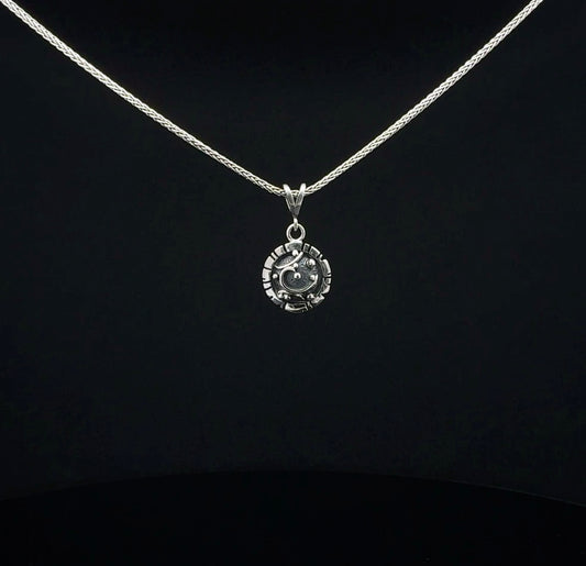 A silver necklace with a small circular pendant featuring a stylized face, displayed against a solid black background.