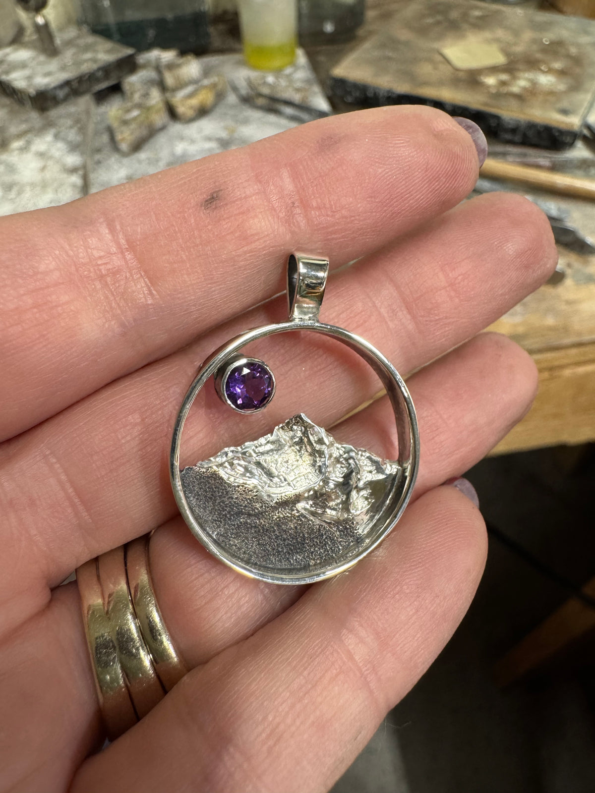 A sterling silver cone-shaped pendant with an engraved mountain range and a purple gemstone at the top, held in someone's hand.