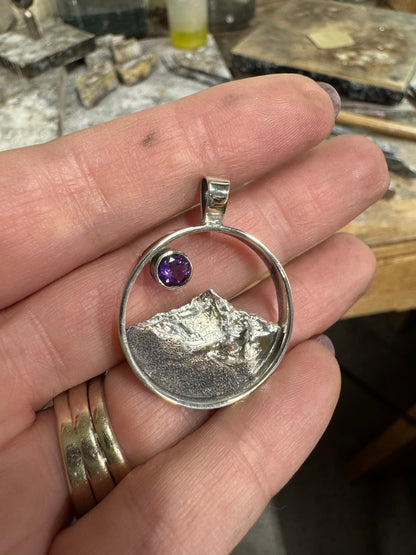 A sterling silver cone-shaped pendant with an engraved mountain range and a purple gemstone at the top, held in someone's hand.
