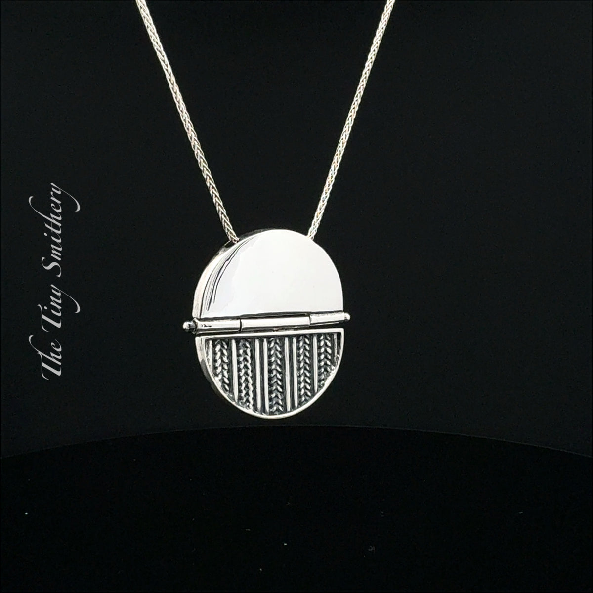 A silver pendant necklace featuring a split circular design, with one half polished and the other half textured with vertical and chevron patterns, suspended from a delicate silver chain against a black background. The brand name 'The Tin Smithery' is visible as a watermark on the left.