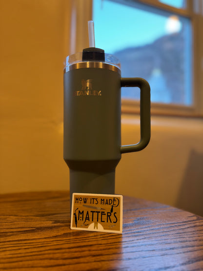 A dark olive green Stanley tumbler with a handle and clear lid, standing on a wooden table with a small sign in front. A window and warm indoor wall are blurred in the background.