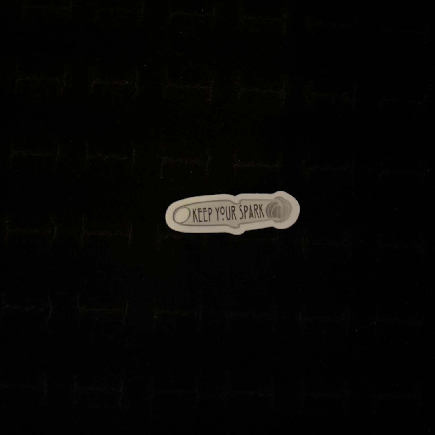 A sticker with the phrase 'Keep Your Spark' printed on it, placed against a dark background.