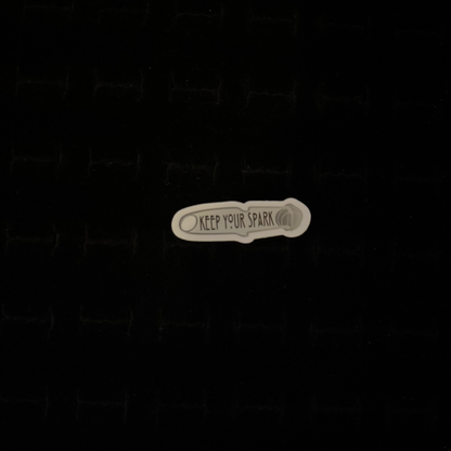 A sticker with the phrase 'Keep Your Spark' printed on it, placed against a dark background.