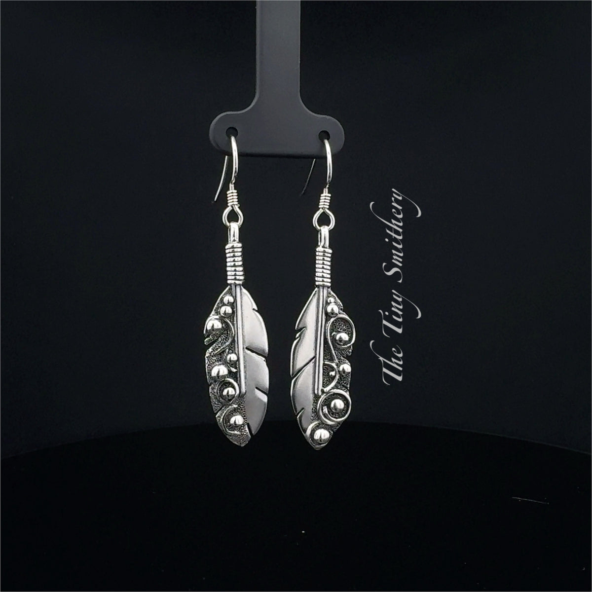 Silver feather-shaped earrings on a black stand with 'The Wire Smith' branding.