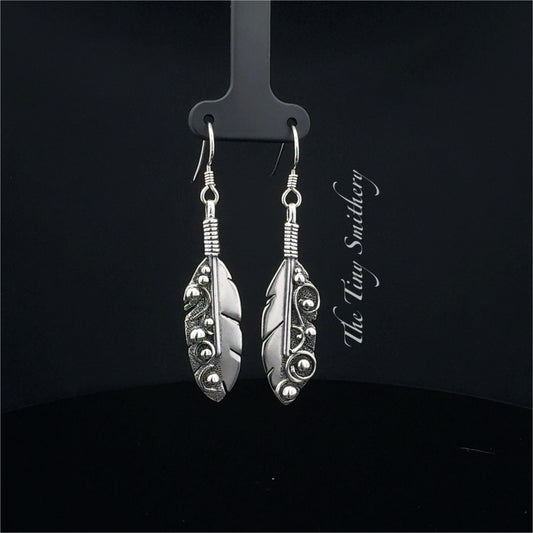 Silver feather-shaped earrings on a black stand with 'The Wire Smith' branding.