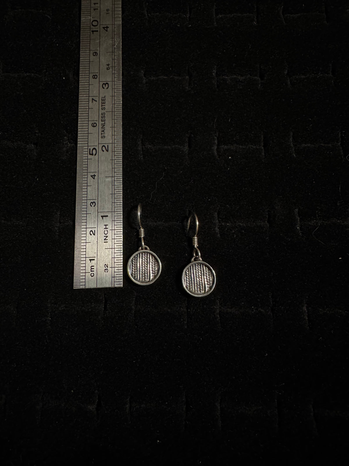Pair of round earrings on a dark textured surface with a ruler for scale.