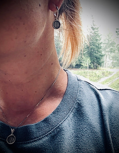 A close-up lifestyle shot of a person wearing a silver-toned circular pendant necklace and matching drop hoop earring with a dark engraved design, set against a blurred outdoor background.