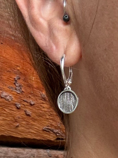A close-up of a woman wearing a delicate silver chain necklace with a round silver pendant and a matching silver hoop earring, set against a background of a wooden wall and her black top.
