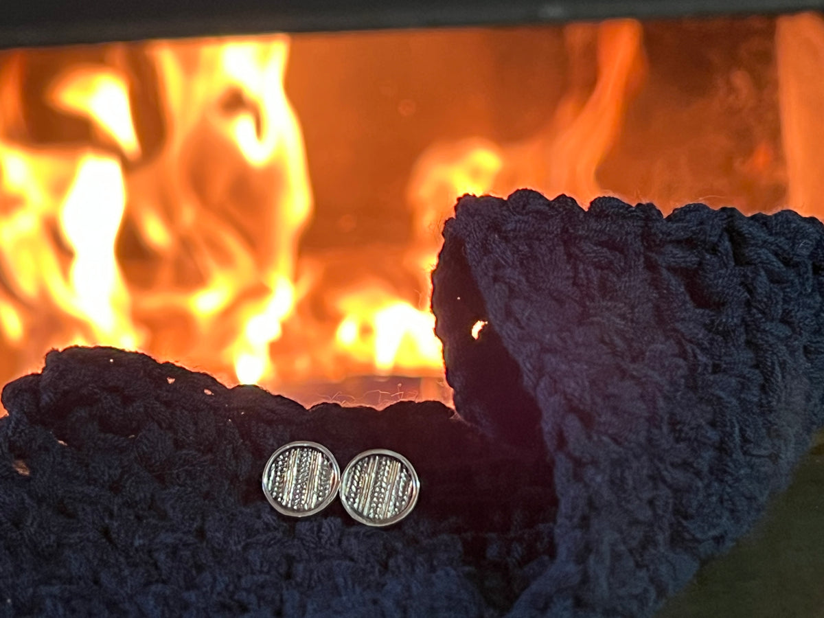 Medium Sweater Post Earrings – lifestyle shot of handcrafted sterling silver knit-pattern disc stud earrings resting on dark knit sweater near fireplace
