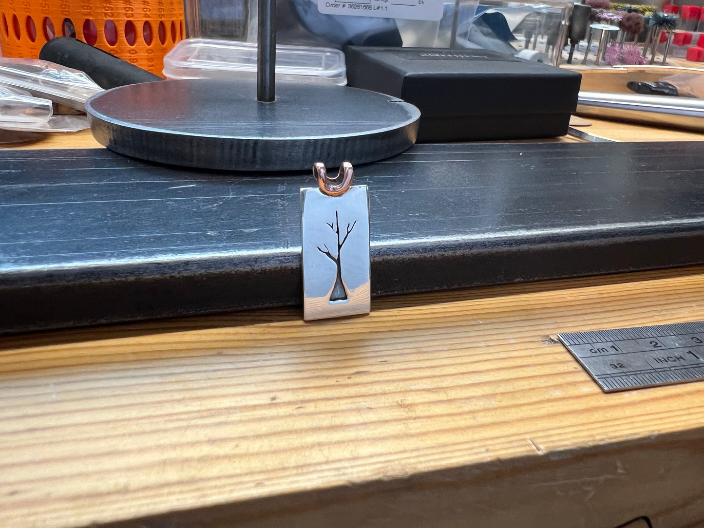 A sterling silver sapling tree pendant with a copper bail, displayed on a wooden surface with tools and equipment in the background.