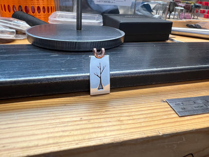 A sterling silver sapling tree pendant with a copper bail, displayed on a wooden surface with tools and equipment in the background.