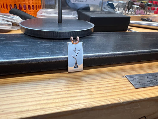 A sterling silver sapling tree pendant with a copper bail, displayed on a wooden surface with tools and equipment in the background.