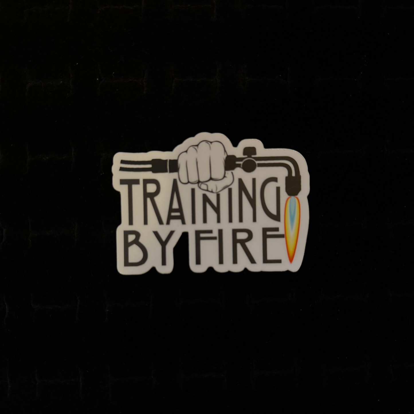 A sticker with a graphic of a torch and the phrase 'TRAINING BY FIRE' on a dark background.
