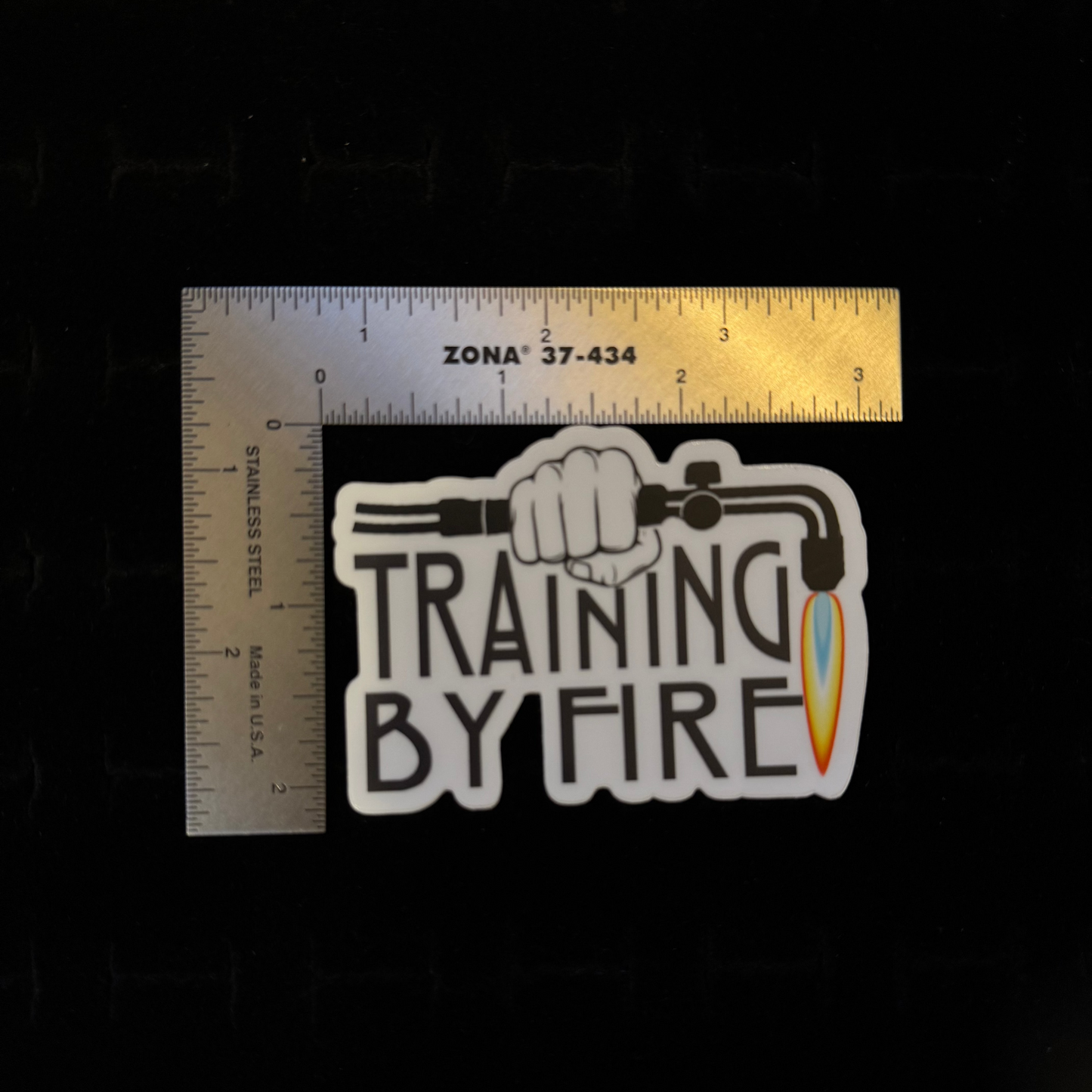 "Training By Fire" sticker – size reference showing white die-cut Training By Fire sticker approximately 2.5 inches wide next to ruler, black background