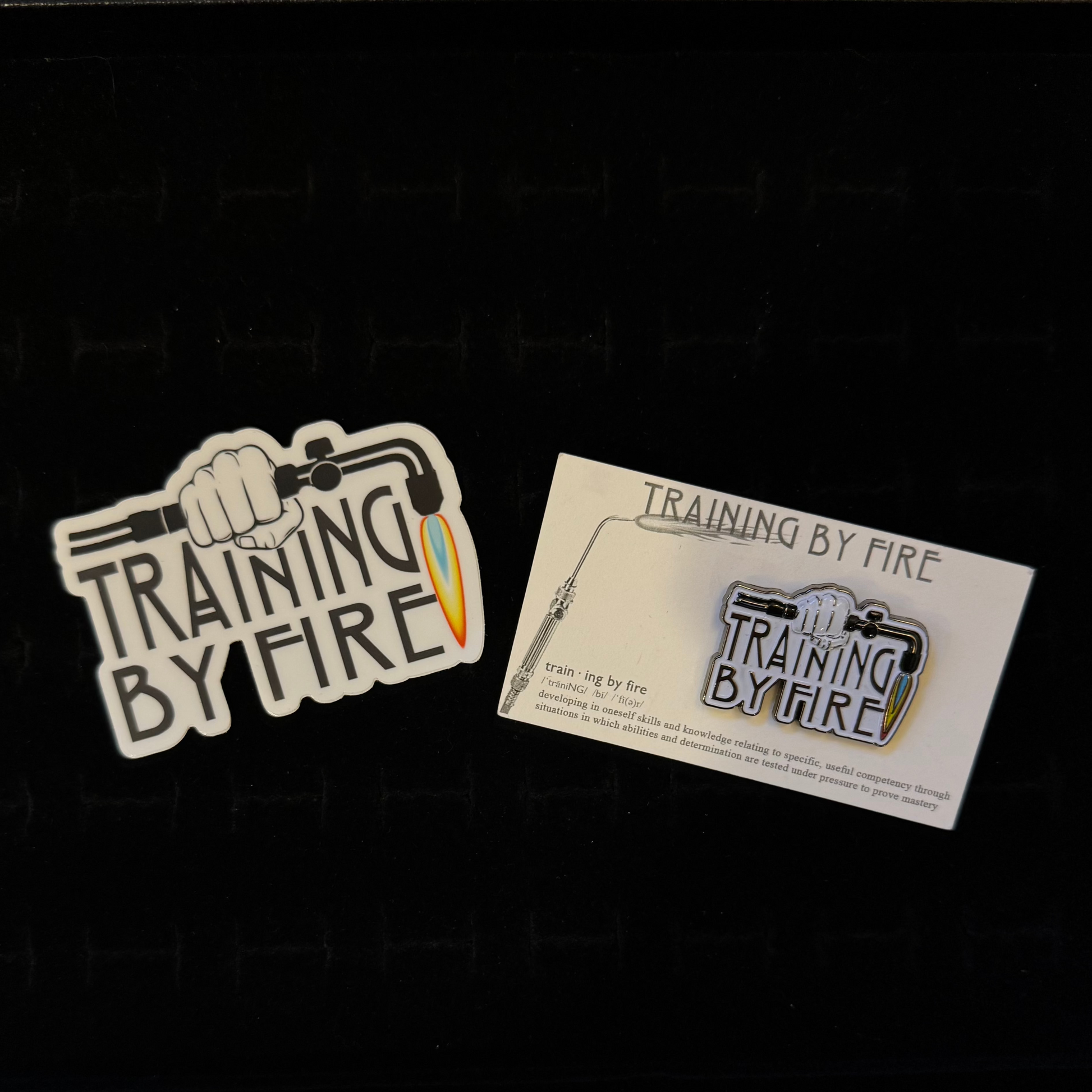
"Training By Fire" sticker – flat lay of white die-cut Training By Fire sticker with torch and fist graphic, black background
