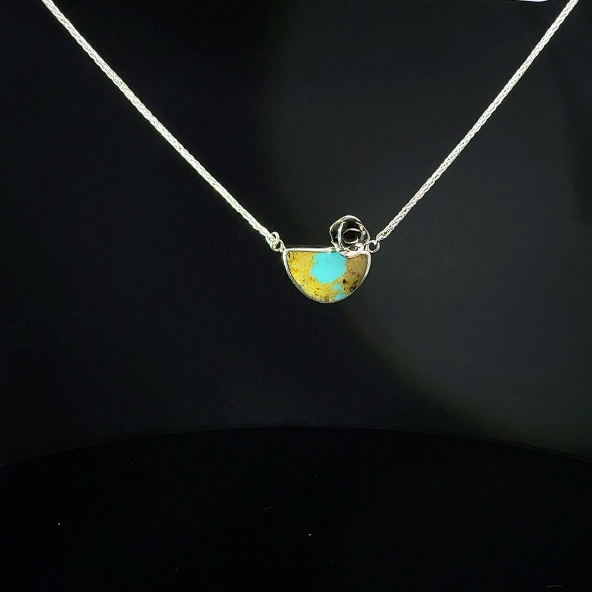 A silver chain necklace with a small semicircle pendant featuring a turquoise blue center and gold-toned outer areas, displayed against a dark black background.