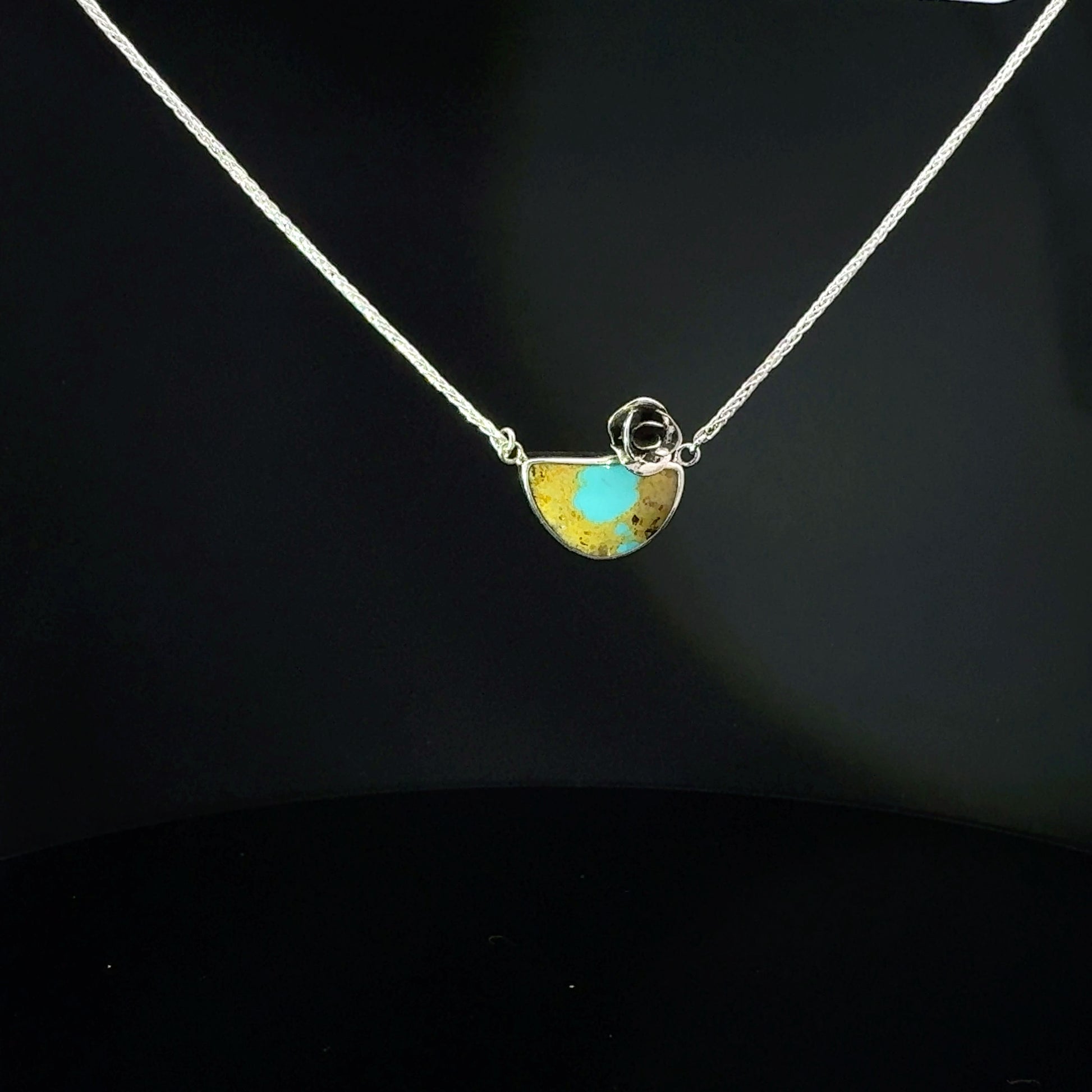 A silver chain necklace with a small semicircle pendant featuring a turquoise blue center and gold-toned outer areas, displayed against a dark black background.