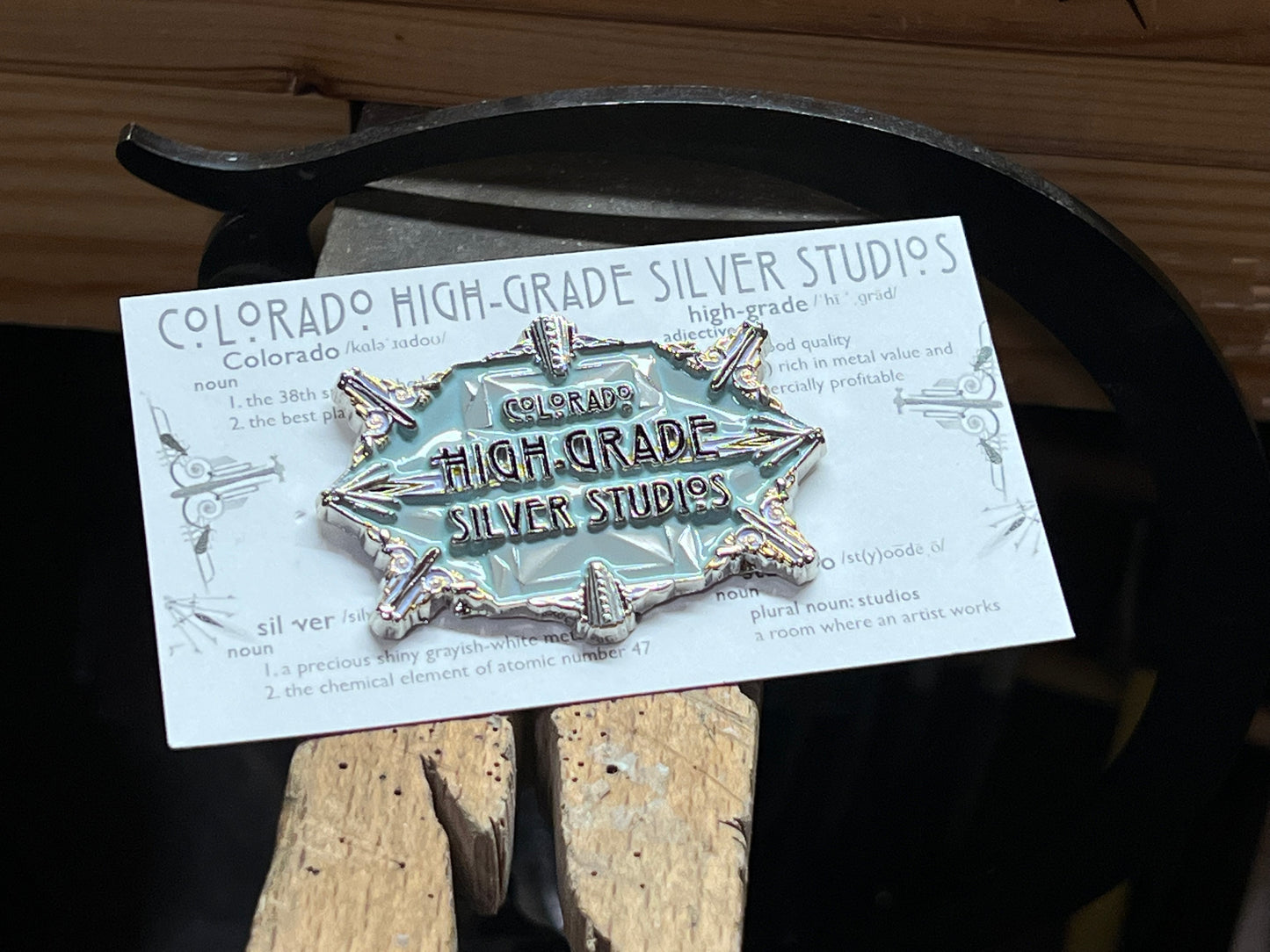 An enamel pin with a faceted gemstone in a setting, surrounded by text that reads 'Colorado High-Grade Silver Studios', placed on a wooden surface.