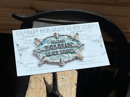An enamel pin with a faceted gemstone in a setting, surrounded by text that reads 'Colorado High-Grade Silver Studios', placed on a wooden surface.