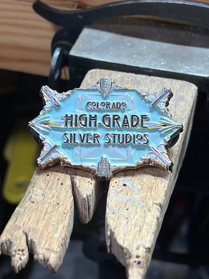 Colorado High-Grade Silver Studios enamel pin – close-up of light blue and silver art deco hard enamel starburst pin with Colorado High-Grade Silver Studios text resting on wooden bench pin