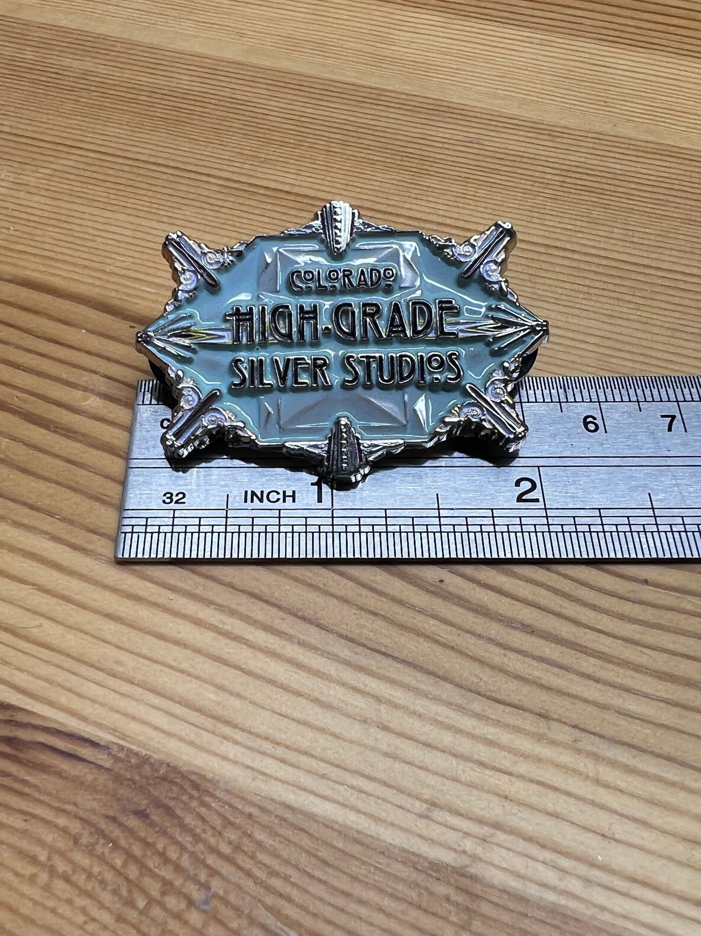 Colorado High-Grade Silver Studios enamel pin – size reference showing large art deco starburst pin approximately 2 inches wide next to ruler, wooden surface