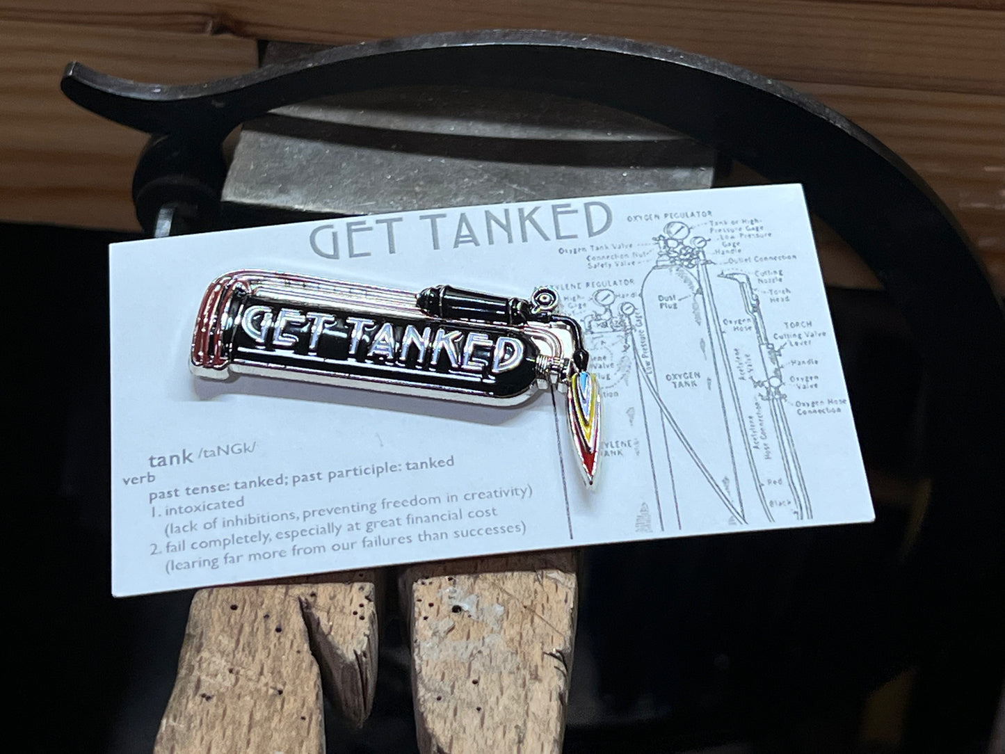 An enamel pin with a jeweler's gas tank design and the words 'GET TANKED' displayed on it, placed on a wooden surface.