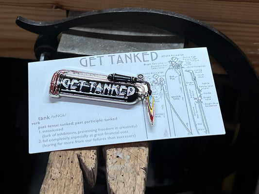 An enamel pin with a jeweler's gas tank design and the words 'GET TANKED' displayed on it, placed on a wooden surface.