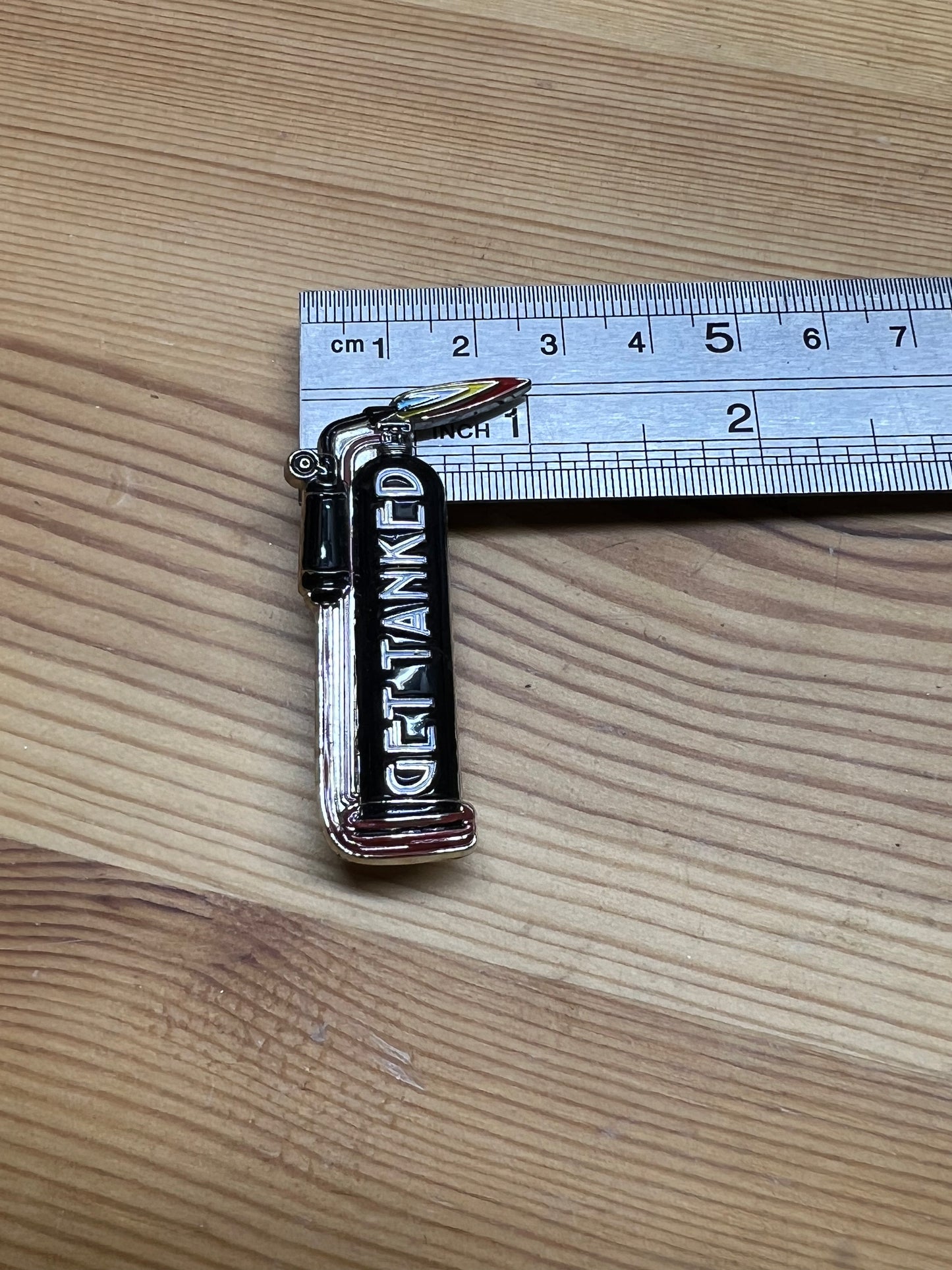 "Get Tanked" enamel pin – size reference showing pin approximately 1.5 inches tall next to ruler, wooden surface