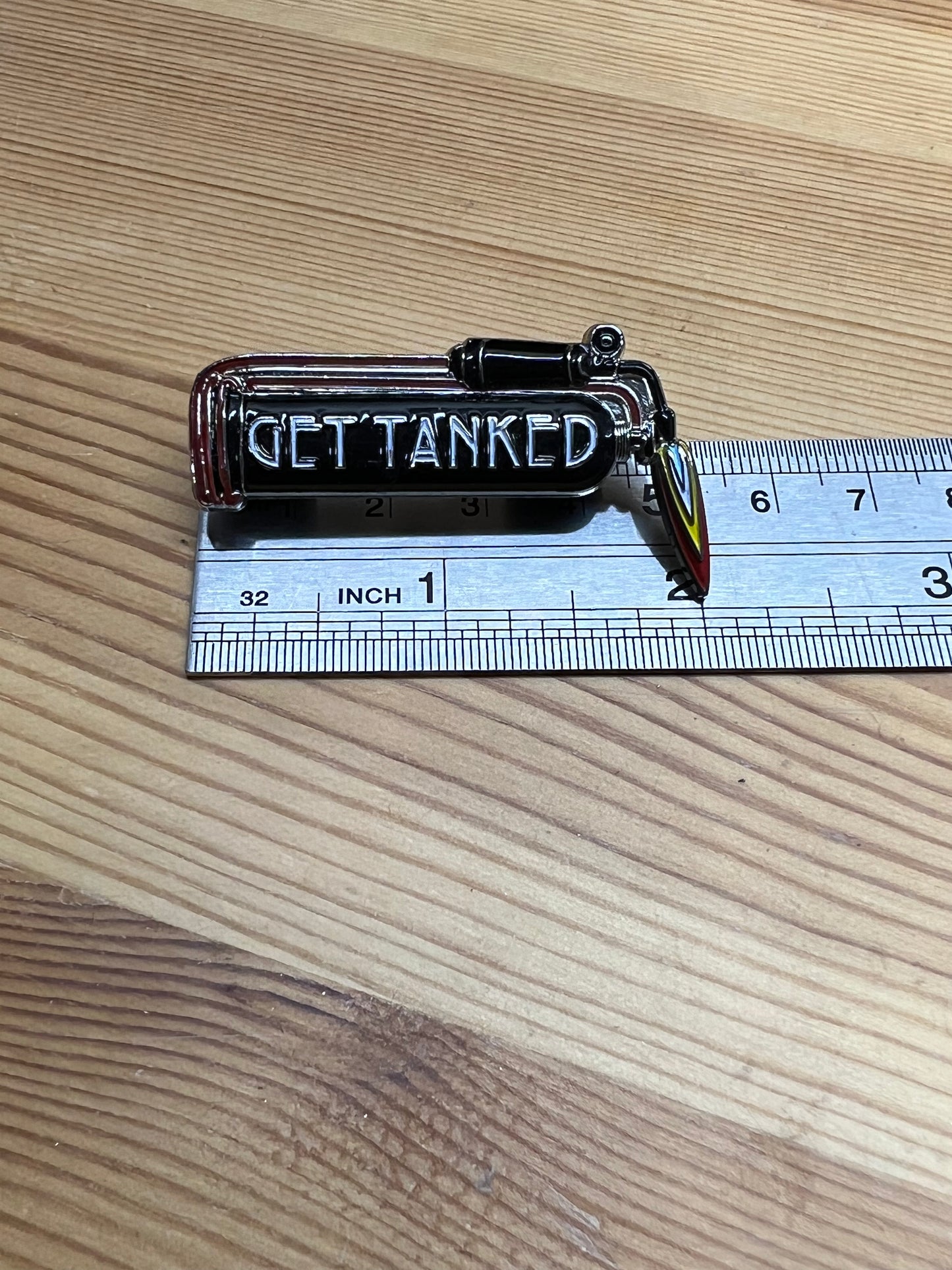 "Get Tanked" enamel pin – horizontal size reference showing pin width approximately 1.5 inches next to ruler, wooden surface
