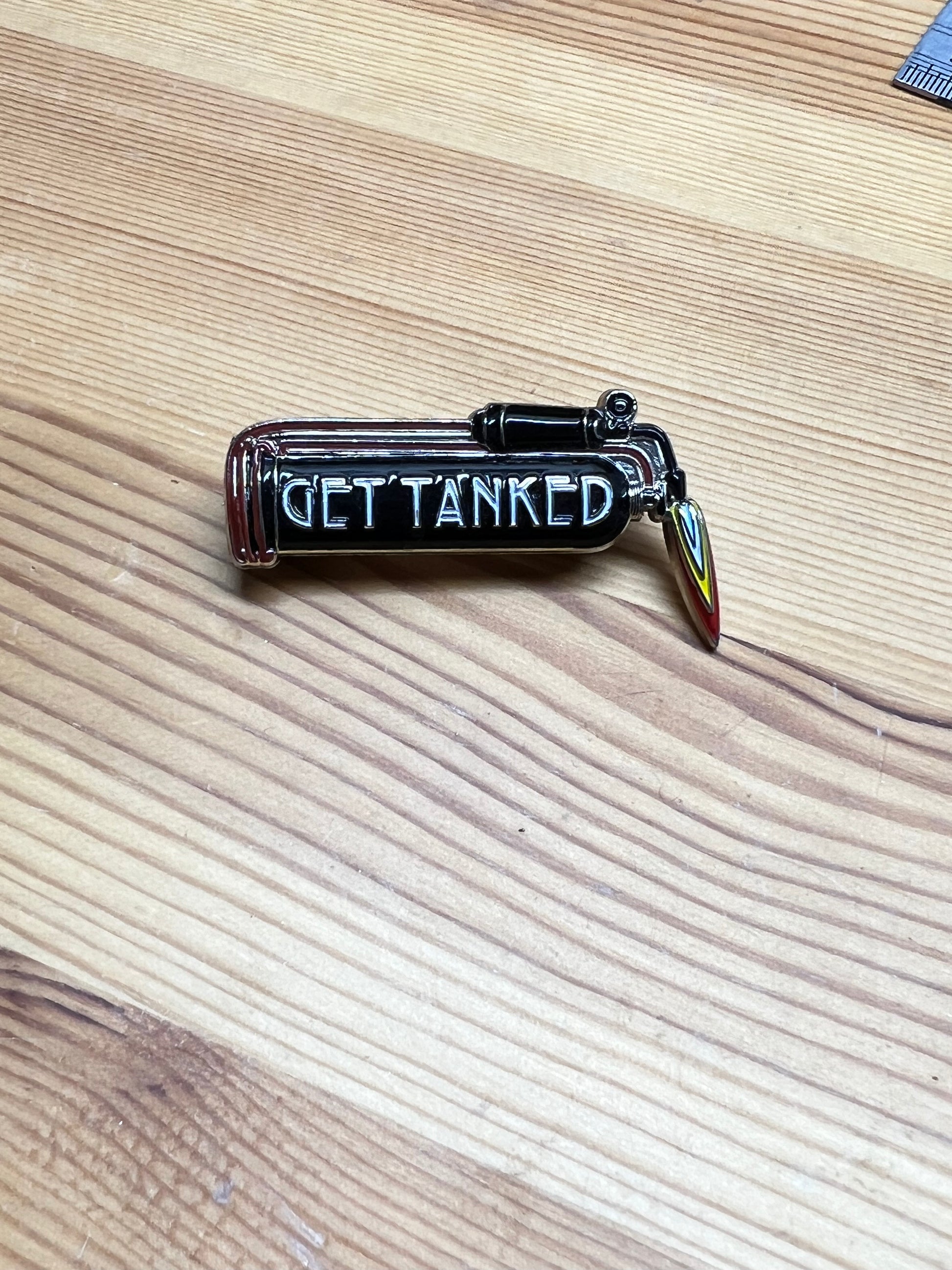 "Get Tanked" enamel pin – front view of black and silver hard enamel pickle tank pin with flame charm, wooden surface