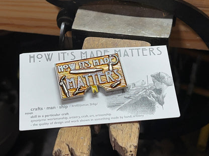 A rectangular enamel pin with a jeweler's bench design and the text 'HOW IT'S MADE MATTERS' on a craftsman's shirt.