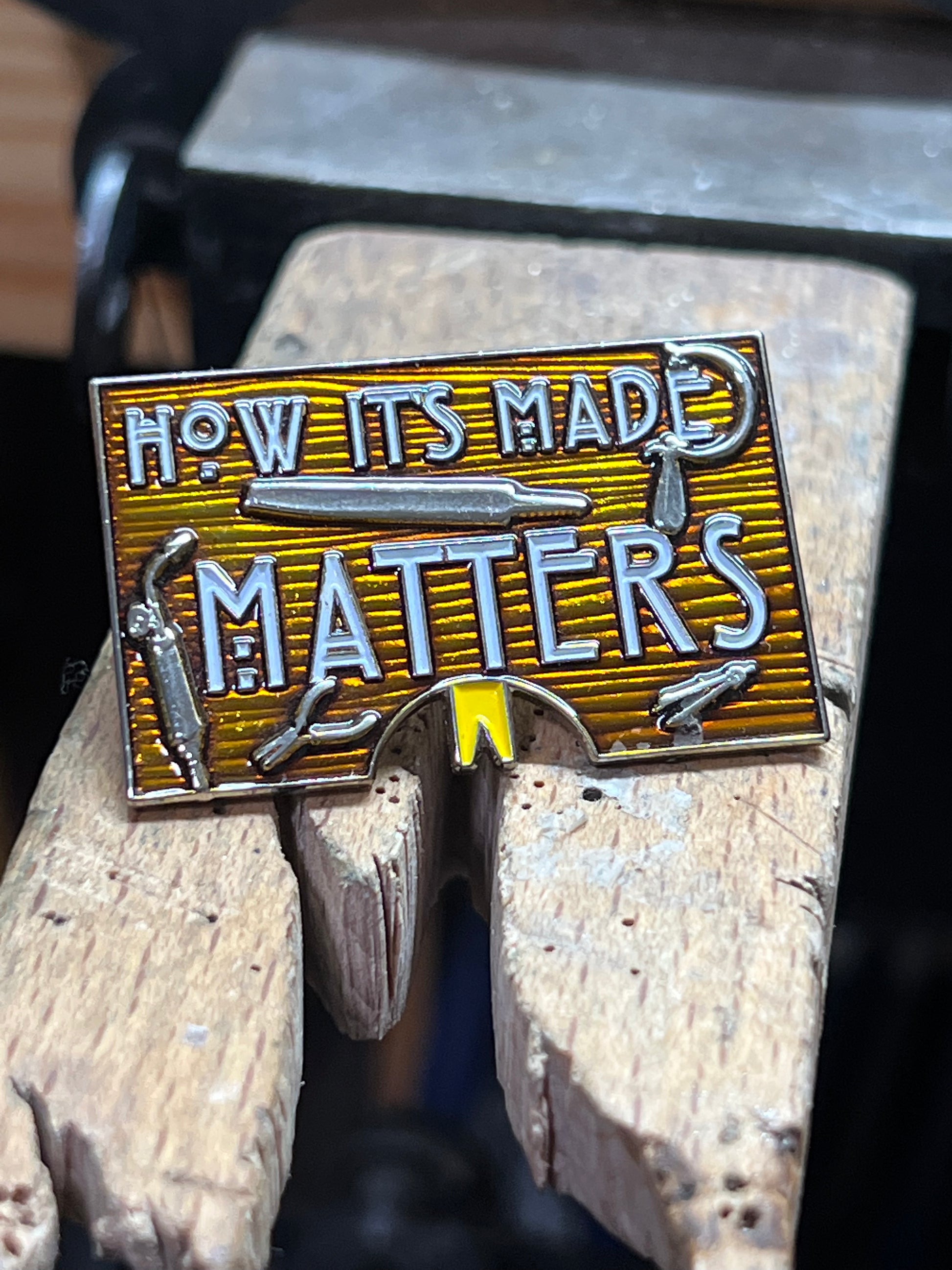 "How It's Made Matters" enamel pin – close-up of gold and silver hard enamel rectangular pin with How Its Made Matters text and jeweler's tools detail, resting on wooden bench pin