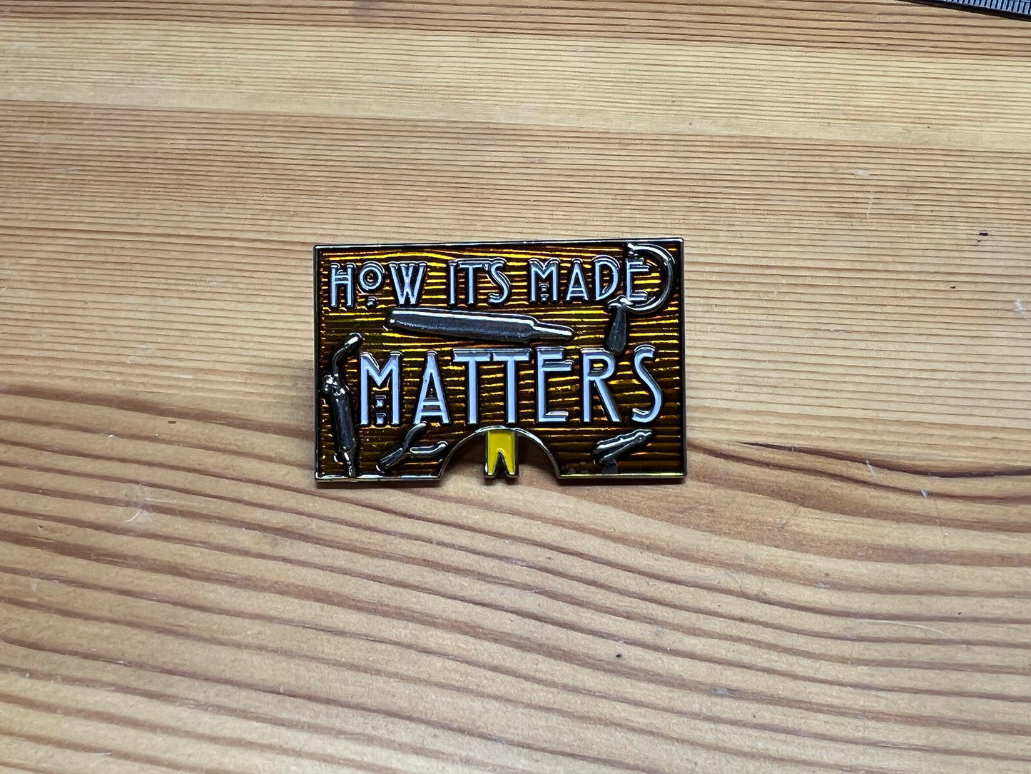 "How It's Made Matters" enamel pin – front view on wooden surface
