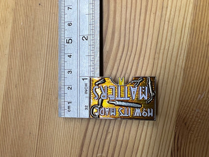 "How It's Made Matters" enamel pin – size reference next to ruler, wooden surface