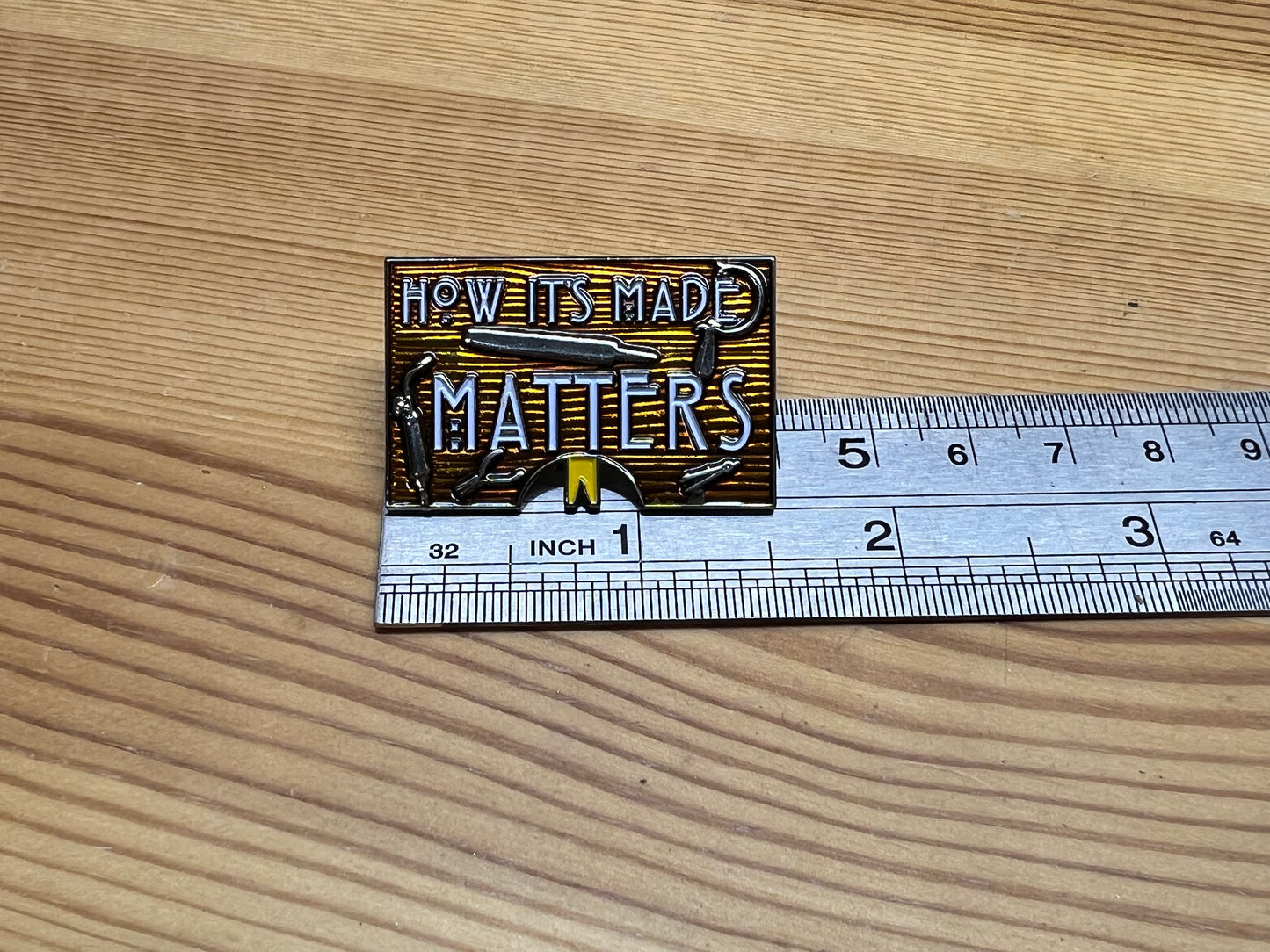 "How It's Made Matters" enamel pin – back view showing butterfly clasp hardware, wooden surface