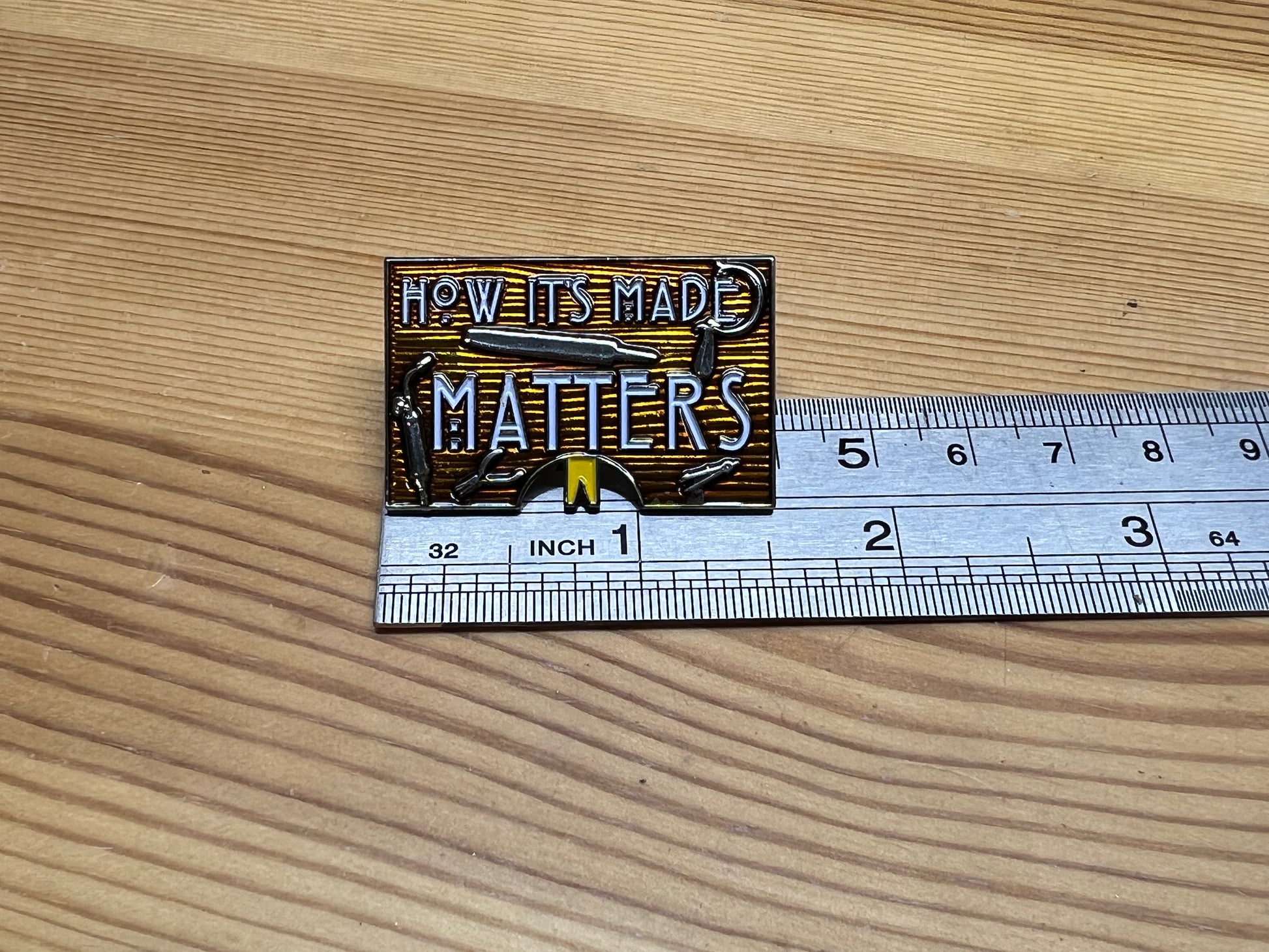 "How It's Made Matters" enamel pin – back view showing butterfly clasp hardware, wooden surface