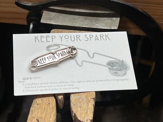 An enamel pin with a jeweler's striker and the words 'Keep Your Spark' displayed on a metal surface with a wood background.