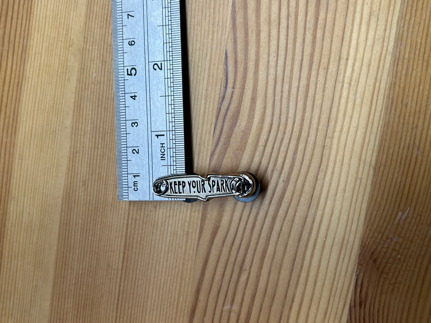 "Keep Your Spark" enamel pin – size reference showing pin approximately 1.5 inches tall next to ruler, wooden surface