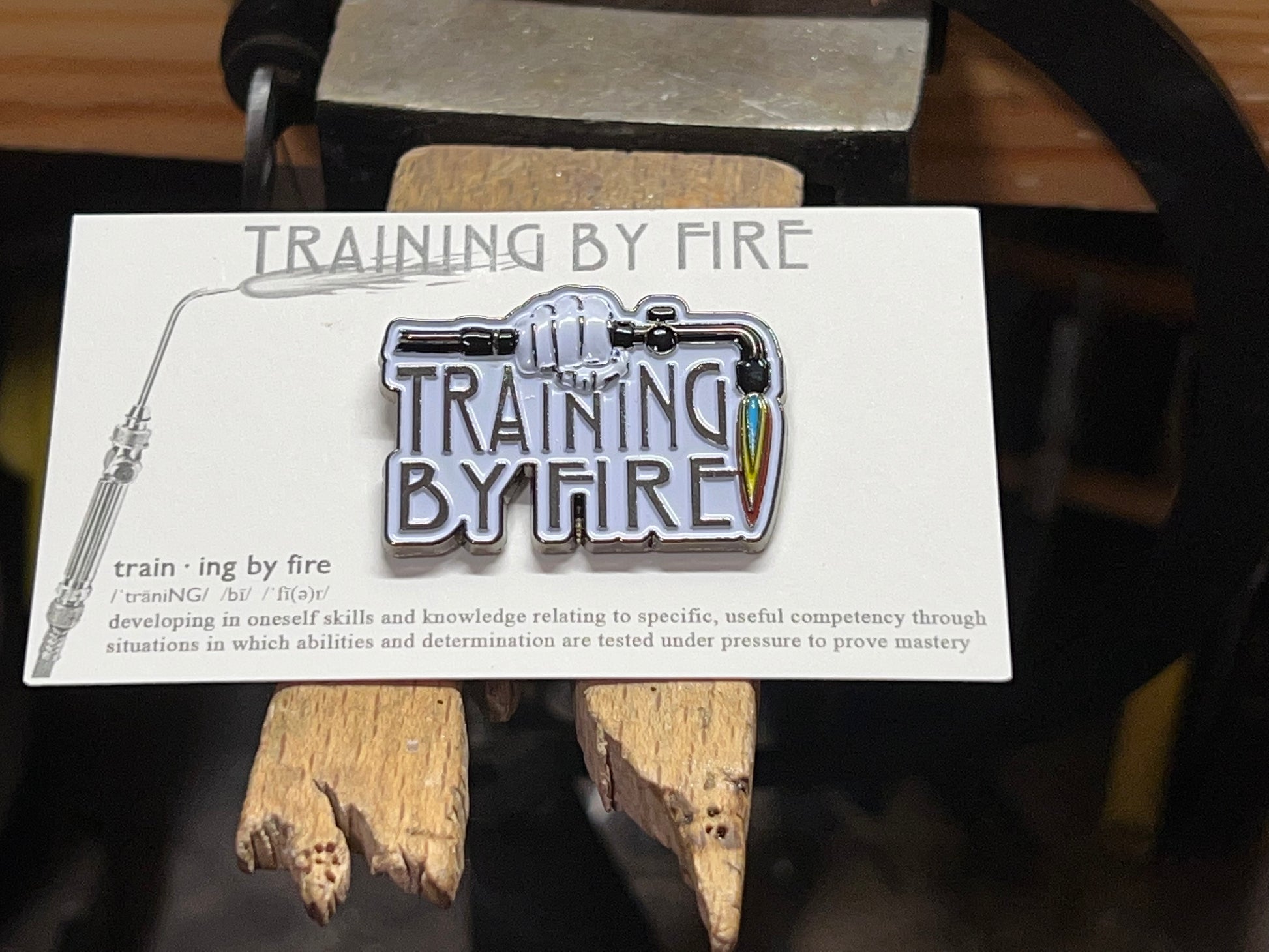 An enamel pin with a jeweler's torch design and the text 'TRAINING BY FIRE' displayed on it, placed on a wooden surface.