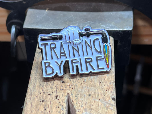 "Training By Fire" enamel pin – close-up of hard enamel black and silver torch pin with Training By Fire lettering, dark background