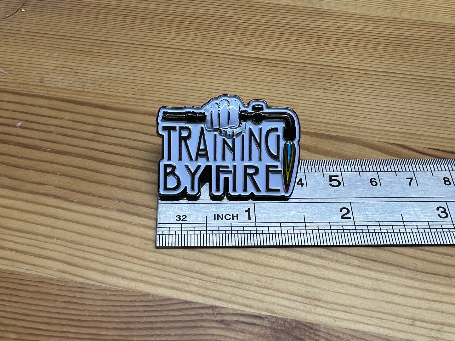"Training By Fire" enamel pin – size reference next to ruler on wooden surface