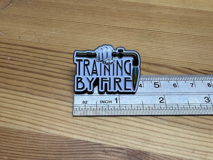 "Training By Fire" enamel pin – size reference next to ruler on wooden surface